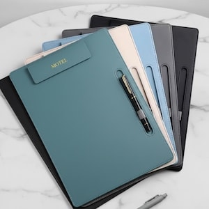 Può includere: Una pila di cartelle portablocco in pelle in vari colori: verde acqua, crema, azzurro, grigio e nero. Ogni cartella portablocco ha un portapenne e la scritta "MOTEL" in rilievo sulla parte superiore. Una penna è appoggiata sulla superficie.