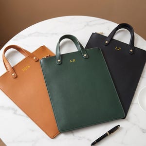 Peut inclure: Trois porte-documents rectangulaires de couleur fauve, vert foncé et noir, chacun doté de deux poignées supérieures, d'une fermeture éclair dorée et d'initiales dorées. Le modèle fauve porte TINA, le vert A.R et le noir L.P en lettres dorées.