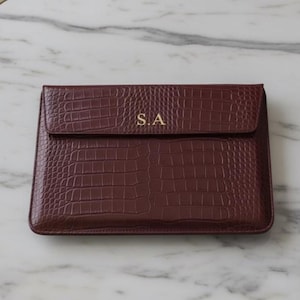 May include: A maroon laptop case with a faux crocodile skin pattern. The initials "S.A" are in gold on the flap. The case is rectangular and rests on a white and grey marble table.