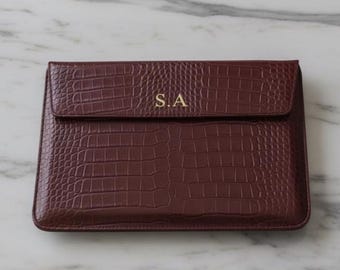 May include: A burgundy laptop sleeve with a crocodile skin texture. The flap closure is embossed with the gold initials "S.A." The sleeve is rectangular and sits on a white and gray marble table.