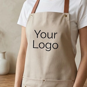 May include: A beige apron featuring the text Your Logo in black lettering. The apron includes brown leather-like neck straps, metallic rivets, and two front pockets, one of which has a small rectangular brown leather-like patch on the right side.