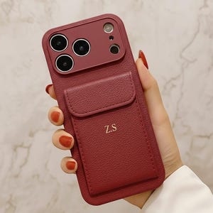 May include: A burgundy phone case with a cardholder and camera cutouts. The case has a textured finish and the initials "Z.S" are embossed on the cardholder. The case is designed to fit a smartphone.