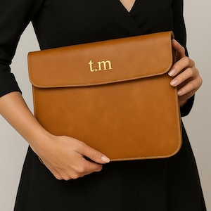 Könnte beinhalten: Eine hellbraune Laptop-Hülle aus Leder mit den goldenen Buchstaben "t.m" auf der Vorderseite. Die Hülle hat einen Klappverschluss und wird von einer Person in einem schwarzen Kleid gehalten.