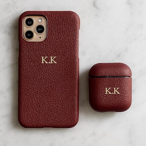 Könnte beinhalten: Ein burgunderfarbenes Leder-Handycase und ein passendes AirPod-Case. Beide Artikel tragen die goldenen Initialen "K.K.". Das Handycase ist für ein Smartphone, und das AirPod-Case ist ein kleiner, abgerundeter Quadrat.
