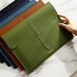 Può includere: Una collezione di porta documenti in pelle in vari colori, tra cui verde, marrone e blu. Il porta documenti verde in primo piano presenta una chiusura con cinturino e le iniziali dorate "K.K.G.G.". I porta documenti sono disposti su una superficie di marmo.