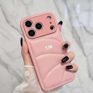 May include: A pink phone case with a quilted design and the initials "T.M" in white. The case has a camera cutout and is held by a hand with black nail polish.
