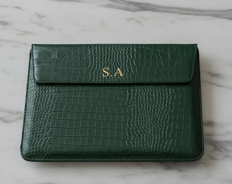 May include: A dark green, faux-leather laptop sleeve with a crocodile skin texture. The flap closure is secured with a magnetic snap. The initials "S.A" are embossed in gold on the front.