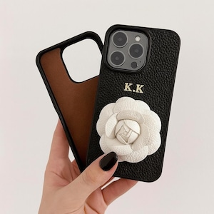 Könnte beinhalten: Schwarze Leder-Handyhülle mit weißem Blumenakzent und den Initialen "K.K.". Die Hülle hat ein braunes Innenfutter und einen schwarzen Rand. Die Handyhülle dient zum Schutz des Telefons.