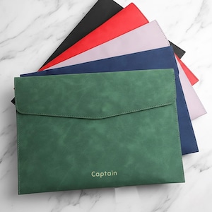 Può includere: Una pila di cartelle colorate: nero, rosso, lavanda, blu navy e verde. La cartella verde, con la parola "Captain" in oro, è in primo piano. Le cartelle sono realizzate in un materiale strutturato.