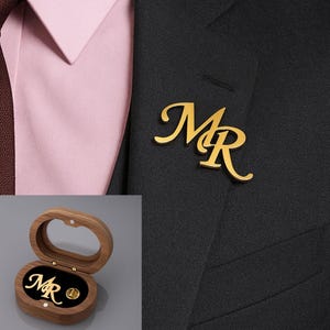 Op de afbeelding: Goudkleurige reversspeld met de letters "MR" in een sierlijk lettertype, gedragen op een zwart colbert. De speld wordt ook getoond in een houten presentatiedoos met een zwart fluwelen interieur. Een roze overhemd en bruine stropdas zijn zichtbaar.