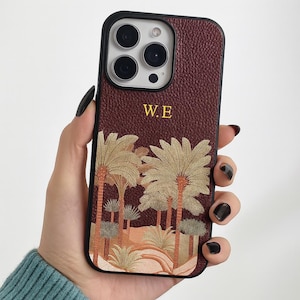 May include: Burgundy leather phone case with a black border. The case features a palm tree design at the bottom and the initials "W.E." in gold lettering at the top.