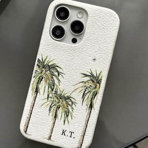 May include: White textured phone case featuring a palm tree design with green and brown details. The case has a camera cutout and the initials "K.T." are printed at the bottom.