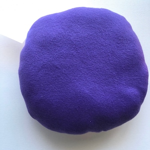 May include: A round, purple, plush pillow. The pillow is made of a soft, fuzzy material.