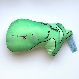 May include: Green plush toy shaped like a cartoon whale with a tongue sticking out. The whale has a smiling face and the words "Fresh Crayons" written on its side.