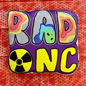 Op de afbeelding: Een paars en geel kussen met de woorden "RAD ONC" in een kleurrijk, cartoonachtig lettertype. Het kussen heeft een geel, zwart en wit cirkelvormig ontwerp in de linkerbenedenhoek.