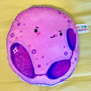 May include: A plush purple cell with a cute cartoon face and two purple organelles. The cell is smiling and has a sparkle on one of the organelles. The plush toy is made by Fresh Crayons.