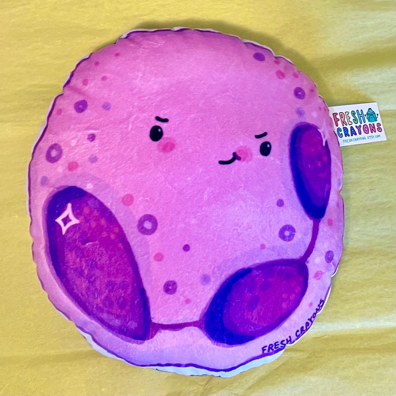May include: A plush purple cell with a cute cartoon face and two purple organelles. The cell is smiling and has a sparkle on one of the organelles. The plush toy is made by Fresh Crayons.
