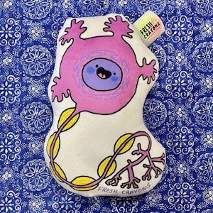 May include: A white plush toy shaped like a neuron, featuring a pink cell body with a smiling face and a purple nucleus. Yellow and pink tendrils extend from the body. The tag reads "Fresh Crayons".