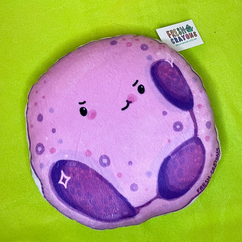 May include: A plush purple and pink cell with a cute cartoon face. The cell has a purple nucleus and a white sparkle. The plush toy is labeled "Fresh Crayons".