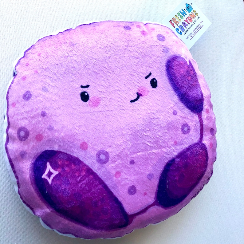 May include: A plush purple cell with a smiling face and two purple organelles. The cell is a cartoon character with a pink blush on its cheeks. The cell is a handmade plush toy from Fresh Crayons.