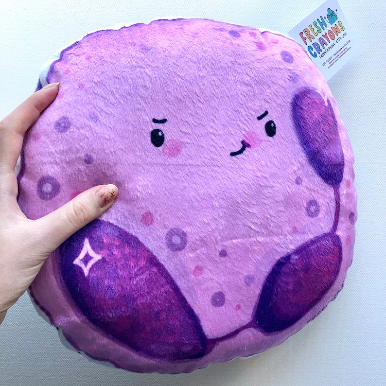 May include: A plush purple and pink cell pillow with a cartoon face and a purple and pink cell structure. The pillow has a white tag that says "FRESH! CRAYONS" with a website address and a copyright symbol.