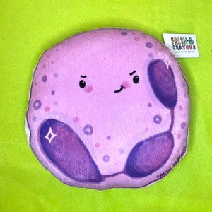May include: A plush purple and white pillow shaped like a cell with a cartoon face. The cell has a nucleus, cytoplasm, and a small diamond shape. The pillow has the text "FRESH CRAYONS" on the bottom.