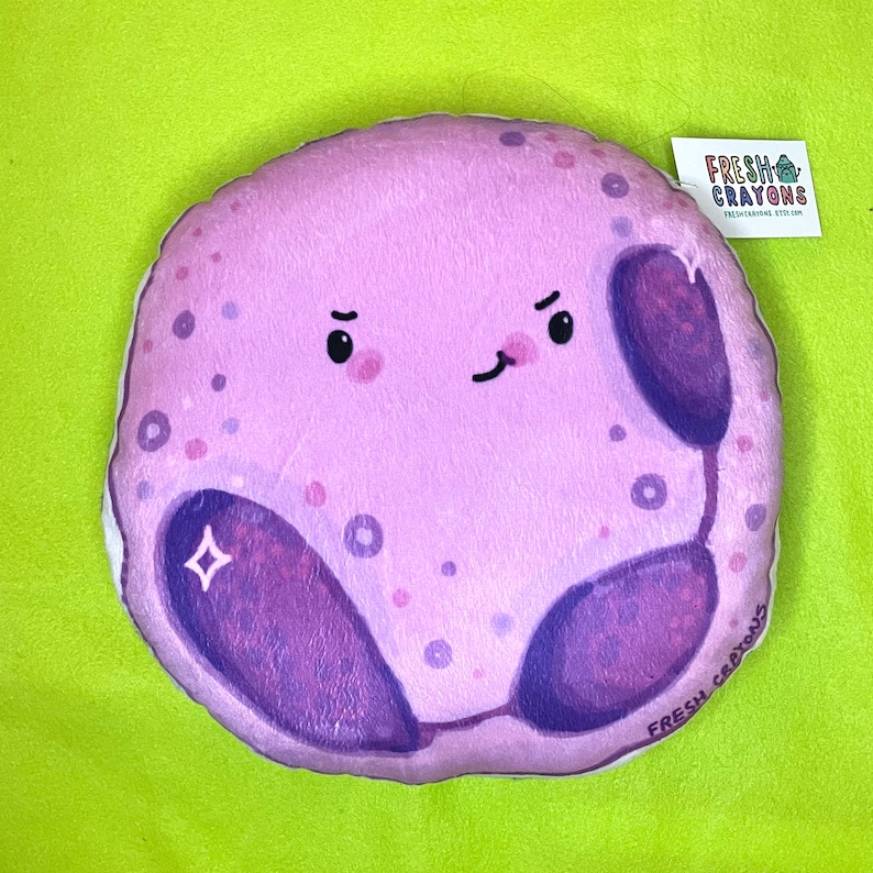May include: A plush purple and white pillow shaped like a cell with a cartoon face. The cell has a nucleus, cytoplasm, and a small diamond shape. The pillow has the text "FRESH CRAYONS" on the bottom.