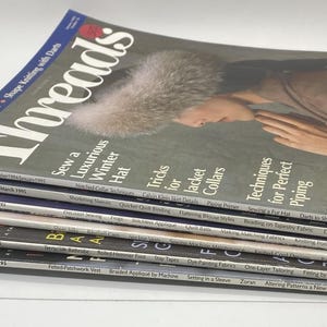 Puede incluir: Una pila de revistas vintage "Threads", con artículos sobre costura y moda. Las portadas muestran imágenes de ropa y técnicas de costura. Las revistas están apiladas en ángulo, con la portada superior mostrando a una mujer con un sombrero de piel.