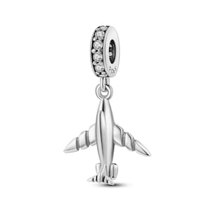 Puede incluir: Un charm de avión plateado con un anillo de piedras transparentes. El avión tiene alas y cola detalladas. El charm está diseñado para ser un colgante o abalorio para una pulsera o collar.
