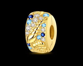 Puede incluir: Un charm dorado con un diseño floral. El charm está adornado con piedras preciosas azules, celeste y transparentes. El charm tiene una abertura circular en un lado.