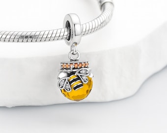 May include: A silver charm with a honey pot design. The charm features a yellow honey pot with a black and yellow bee, and orange and clear accents. The charm is attached to a silver bracelet.