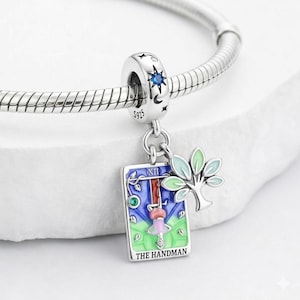 Puede incluir: Un charm de plata con un diseño de estrella y luna azules, sujeto a una pulsera. El charm presenta una colorida carta del tarot con el texto "THE HANDMAN" y un pequeño árbol. La carta del tarot tiene una figura colgada boca abajo.