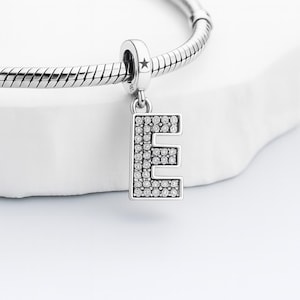 Könnte beinhalten: Ein silbernes Charm-Armband mit einem Anhänger in Form des Buchstabens "E". Der Buchstabe ist mit kleinen, klaren Kristallen besetzt. Der Anhänger ist mit einem silbernen Armband mit sternförmigem Akzent verbunden.