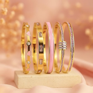 May include: A selection of gold-coloured bangles with diverse designs. One bangle has pink hearts and clear stones, while others feature rectangular clear stones or pink enamel. The last bangle is decorated with pink gemstones.