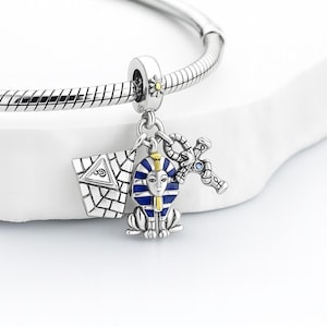 May include: Silver charm bracelet with three dangling charms. Charms include a silver and blue pharaoh, a silver ankh, and a silver and white pyramid. The bracelet is on a white surface.