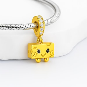 Peut inclure: Un charm doré avec un design de bloc de fromage. Le charm a un visage souriant avec des yeux noirs et une bouche incurvée. Le charm est attaché à un anneau doré avec des pierres claires. Le charm est suspendu à un bracelet argenté.