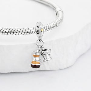 May include: Silver charm with a coffee theme. The charm features a coffee maker and a coffee cup with the word "Coffee" on it. The charm is attached to a silver bracelet.