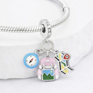 Puede incluir: Una pulsera de plata con un charm temático de viaje. El charm presenta una brújula, una mochila rosa con un diseño azul y verde y un mapa colorido con un alfiler rojo. El charm está unido a una pulsera de plata.