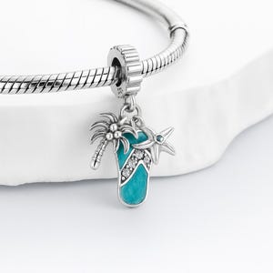 Op de afbeelding: Een zilveren bedel met een tropisch thema. De bedel heeft een turquoise teenslipper met heldere kristallen, een palmboom en een zeester. De bedel is bevestigd aan een zilveren armband.