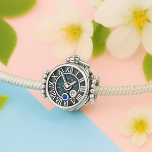 Puede incluir: Un charm de color plateado con un diseño de esfera de reloj. El reloj presenta números romanos, un centro de color turquesa y una pequeña gema azul y transparente. El charm está en una cadena plateada, con flores blancas y hojas verdes en el fondo.