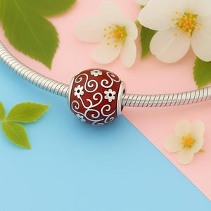 Puede incluir: Una cuenta roja y plateada con un diseño floral. La cuenta es parte de una pulsera o collar, con una cadena plateada visible. Flores blancas y hojas verdes en el fondo, sobre un fondo rosa y azul.