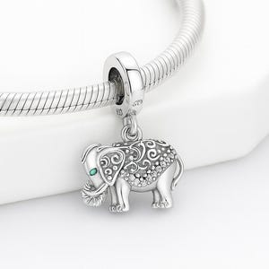 Könnte beinhalten: Ein silberner Elefanten-Charm mit aufwendigen Schnörkeln und einem kleinen grünen Edelsteinauge. Der Charm hängt an einem silbernen Armband. Der Körper des Elefanten ist mit kleinen, funkelnden Akzenten verziert.