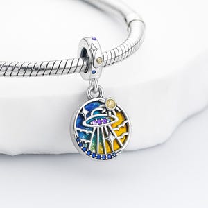 Peut inclure: Un charm en argent avec un pendentif rond coloré représentant une scène d'OVNI. Le pendentif présente un ciel bleu, des nuages jaunes et un OVNI violet et argenté. Le charm est attaché à un bracelet en argent.