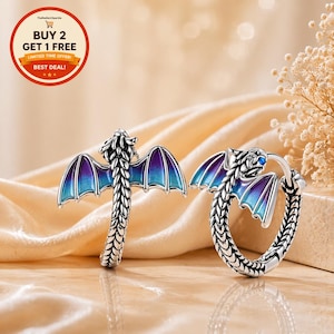 May include: Two silver dragon rings with blue and purple enamel wings. The rings have detailed scales and a dragon head with a blue gem eye. The rings are displayed on a satin surface.