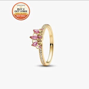 May include: A gold-colored ring with a crown design. The ring features pink gemstone accents and a band embellished with small, clear stones. The ring is set against a white background.