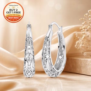 May include: A pair of silver-colored hoop earrings with an intricate, openwork design. The earrings have a detailed, floral-inspired pattern and a polished finish. The image also includes a promotional graphic with text.