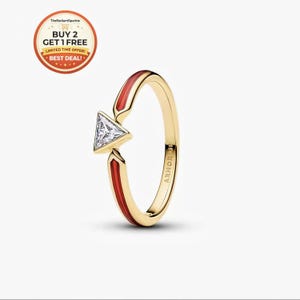May include: A gold ring with a triangular, clear gemstone. The ring features red enamel accents and the text "ARMOR UP" on the band. The ring is set against a white background.