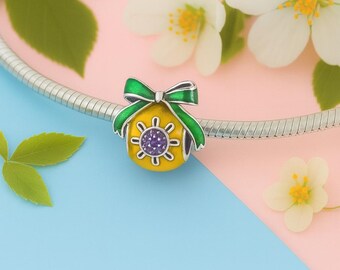 May include: A yellow charm with a green bow and a purple center. The charm is on a silver chain, with white flowers and green leaves in the background. The charm has a floral design.