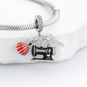 May include: A silver charm bracelet with a sewing-themed charm. The charm features a black sewing machine, a red and white dress form, and silver scissors. The charm is accented with clear crystals.