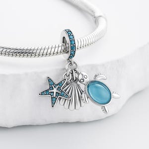 May include: A silver charm bracelet with a sea life theme. The charm features a starfish, seashell, and a turtle, all with blue accents. The charm is attached to a silver bracelet.